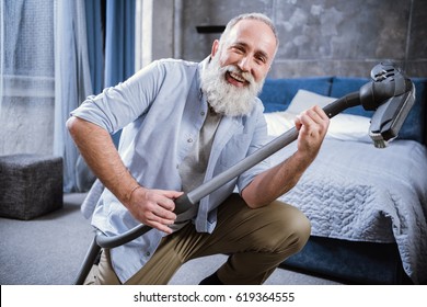 Bearded Senior Man Having Fun With Vacuum Cleaner And Smiling At Camera