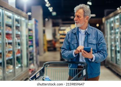 Bearded Senior Man With Shopping Trolley Standing Between Shelves With Food In Grocery Store And Using Phone