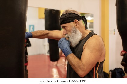 Bearded Senior Man Trains On A Punching Bag. Serious Older Man Working Out With Punching Bag At Boxing Hall. Sport Training Concept.