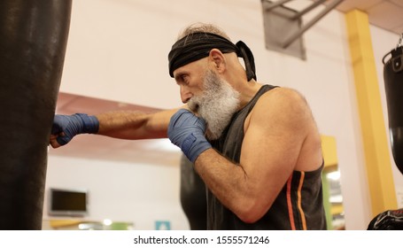 Bearded Senior Man Trains On A Punching Bag. Serious Older Man Working Out With Punching Bag At Boxing Hall. Sport Training Concept.