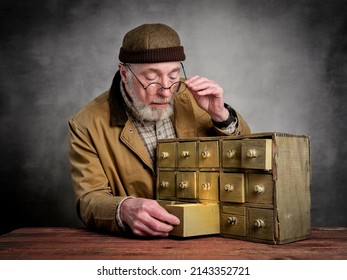 Bearded Senior Man Wearing Glasses And Docker Cap Is Searching For The Light In A Antique Apothecary Cabinet