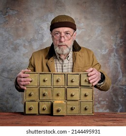 Bearded Senior Man Wearing Glasses And Docker Cap Is Searching For The Light In An Antique Apothecary Drawer Cabinet