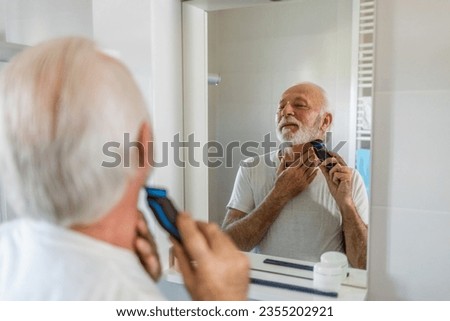 Bearded senior man in white shirt trimming his beard with a trimmer at home.