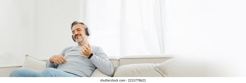 Bearded Senior Mature Adult Man Sitting On Sofa Listening To Music With Headphones And Playing An Air Guitar, Lifestyle Concept, Relaxation At Home. Panoramic, Banner.