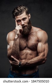 Bearded and shirtless guy with modern haircut and huge biceps posing with finger to his head in dark background.