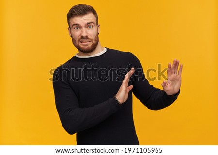 Bearded shocked guy, skeptical looking man with brunette hair. Has piercing. Wearing black sweater. Pushes arms away, reject gesture. Watching at the camera, isolated over yellow background