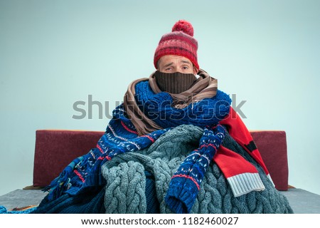 Bearded sick man with flue sitting on sofa at home or studio covered with knitted warm clothes. Illness, influenza, pain concept. Relaxation at Home. Healthcare Concepts.