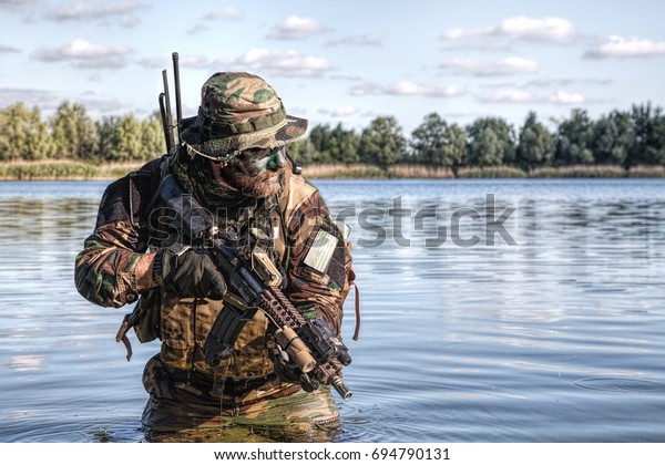 Bearded soldier of special forces in action\
during river raid in the jungle terrain. He is waist deep in the\
water and mud and ready to meet enemy, survive and fight in\
agressive hostile\
environment
