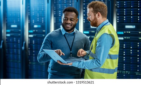 Bearded IT Specialist in Glasses and High Visibility Vest with a Laptop Computer and Black Technician Colleague Talking in Data Center while Standing Next to Server Racks. Running Diagnostics.
