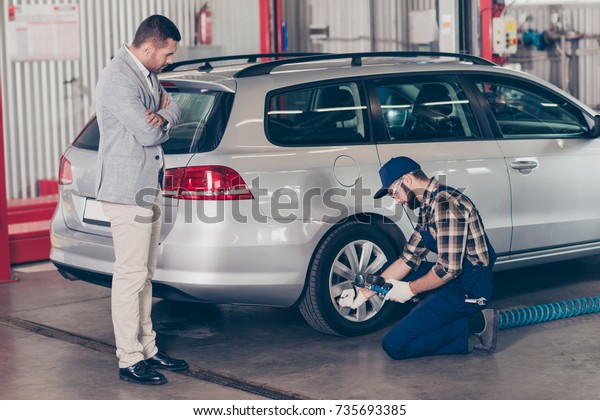 Bearded specialist technician expert in protective\
glasses and blue uniform is analyzing tire pressure of silver car\
on hardware, businessman in classy suit is standing near with\
crossed hands