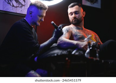 bearded tattoo artist working at his studio tattooing sleeve on the arm of his male client. Man getting tattooed by professional tattooist.