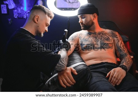 bearded tattoo artist working at his studio tattooing sleeve on the arm of his male client. Man getting tattooed by professional tattooist.