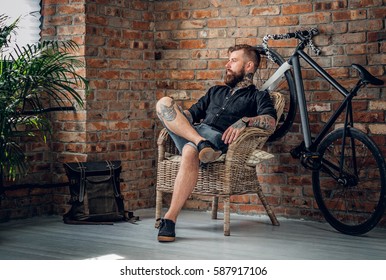 The bearded tattooed male sits on a chair in a room with single speed bicycle on background. - Powered by Shutterstock