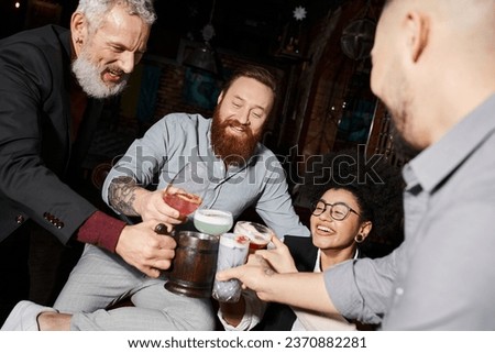 bearded tattooed men and african american woman clinking glasses in cocktail bar, after work fun