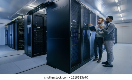 Bearded IT Technician in Glasses with a Laptop Computer and Black Male Engineer Colleague are Talking in Data Center while Working Next to Server Racks. Running Diagnostics or Doing Maintenance Work.