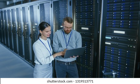 Bearded IT Technician in Glasses with a Laptop Computer and Beautiful Young Engineer Colleague are Talking in Data Center while Working Next to Server Racks. Running Diagnostics or Doing Maintenance  - Powered by Shutterstock
