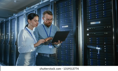 Bearded IT Technician in Glasses with a Laptop Computer and Beautiful Young Engineer Colleague are Talking in Data Center while Working Next to Server Racks. Running Diagnostics or Doing Maintenance 