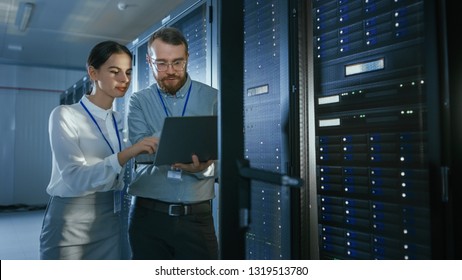 Bearded IT Technician in Glasses with a Laptop Computer and Beautiful Young Engineer Colleague are Talking in Data Center while Working Next to Server Racks. Running Diagnostics or Doing Maintenance 
