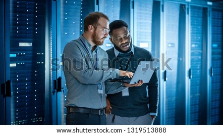 Bearded IT Technician in Glasses with Laptop Computer and Black Male Engineer Colleague are Using Laptop in Data Center while Working Next to Server Racks Running Diagnostics or Doing Maintenance Work