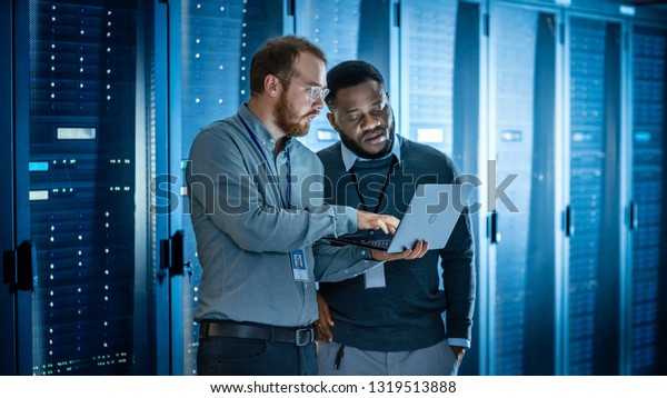 Bearded IT Technician in Glasses with Laptop
Computer and Black Male Engineer Colleague are Using Laptop in Data
Center while Working Next to Server Racks Running Diagnostics or
Doing Maintenance Work