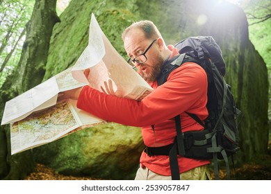 Bearded tourist man looks puzzled, studies map while stands in front of large rock in dense forest. Traveler with grey backpack and glasses, trying to figure out his direction. - Powered by Shutterstock