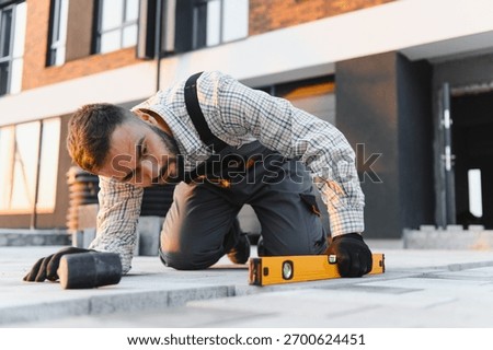 Bearded worker installing paving slabs on ground. Man uses a spirit level for precise and accurate work on construction site