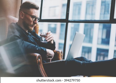 Bearded Young Businessman Working On Modern Loft Office. Man Sitting In Vintage Chair, Holding Glass Water. Using Contemporary Notebook, Panoramic Windows Background. Horizontal, Film Effect. 