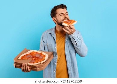 Bearded Young Guy Eating Slice Of Pizza Standing Posing With Box Delivered From Pizzeria Over Blue Studio Background. Junk Food Eater Enjoying Unhealthy Cheat Meal. Male Nutrition And Overeating - Powered by Shutterstock