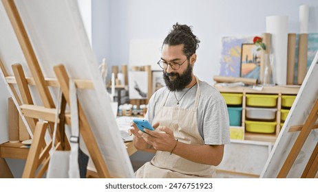 A bearded young man in an apron uses a smartphone in an art studio surrounded by easels and paintings. - Powered by Shutterstock