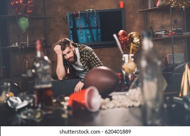 Bearded Young Man With Hangover Sitting On Couch In Messy Room After Party 