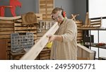 A bearded young man inspects a wooden plank in a well-organized carpentry workshop, wearing protective eyewear and earmuffs.