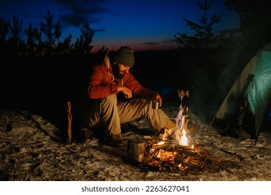 A bearded young man makes delicious coffee on a bonfire while enjoying the sunset in a winter forest. The concept of survival and hiking in the wild. - Powered by Shutterstock