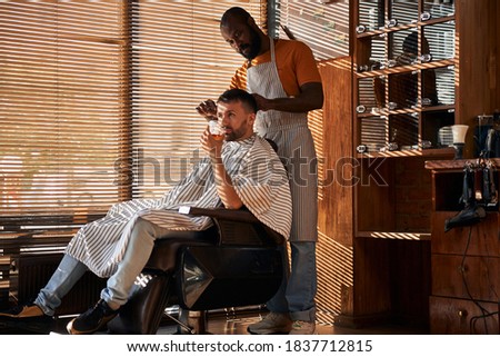Bearded young man sitting in barber chair and drinking whisky while getting haircut in barbershop