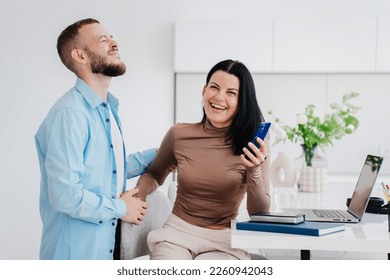 Bearded young man stands eyes closed upset, guy forgot about anniversary holds wife’s hand. Brunette businesswoman smiles wide holds phone shows calendar on phones screen to husband. Happy couple home - Powered by Shutterstock