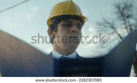 Beardless Construction Engineer or Contractor in a Helmet and Suit Examining Blueprints in a Blue Folder and Getting Frustrated over a Faulty Detail