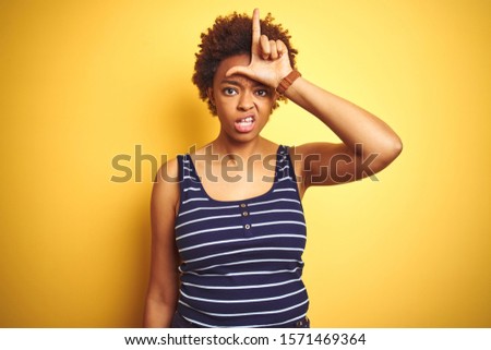 Beauitul african american woman wearing summer t-shirt over isolated yellow background making fun of people with fingers on forehead doing loser gesture mocking and insulting.