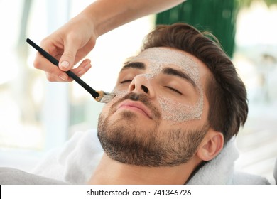 Beautician applying scrub onto young man's face in spa salon