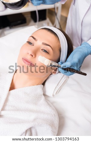 beautician beginning to apply cosmetic mask with cosmetic brush on woman face at beauty salon