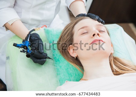 Beautician cosmetologist applying permanent makeup on girl's face. Using tattoo machine. Brow correction in beauty salon