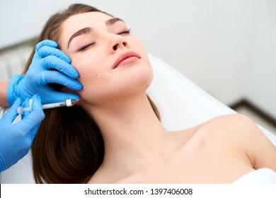 Beautician doctor with filler syringe making injection to jowls. Masseter lines reduction and face contouring therapy. Anti-aging treatment and face lift in cosmetology clinic. Patient lying on chair