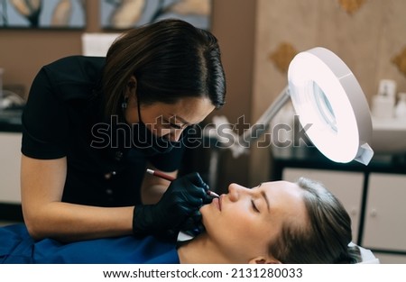 Beautician drawing contour on lips of caucasian girl in beauty salon. Permanent makeup lips procedure. Asian female master wearing gloves and medical mask. Woman client with closd eyes. Beauty concept