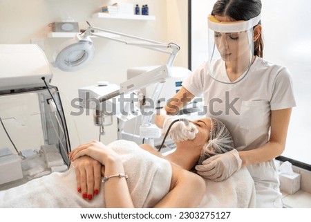 Beautician with face shield performing cosmetology procedures using radio frequency beauty machine for skin care in modern beauty salon. ストックフォト © 