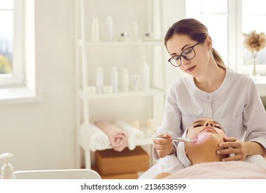 Beautician In Glasses Applying Pink Clay Mask On Woman's Face. Calm Beautiful Young Lady Relaxing And Enjoying Facial Procedures On Spa Day In Modern Beauty Salon Or Spa Center. Copy Space Background