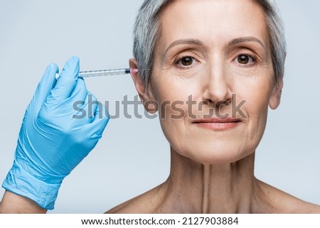 beautician in latex glove holding syringe with beauty injection near cheerful mature woman isolated on grey