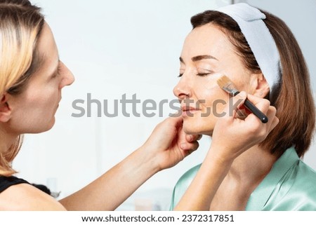Beautician makeup artist putting prime foundation cream with brush on the face of middle aged woman wearing headband