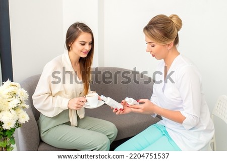 Beautician offering product for the young woman holding white plastic bottles with a cream sitting on the sofa