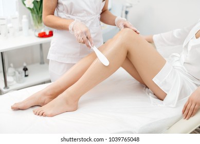 The beautician is preparing for depilation and applying the cream with wax stick on the beautiful female legs. Beauty salon.