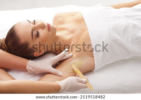 Beautician is removing hair from young female armpits with hot wax. Woman has a beauty treament procedure. Depilation, epilation, skin and health care concepts.
