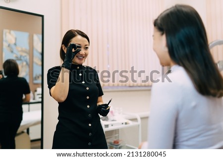 Beautician showing fingers gesture at girl client in beauty salon. Semi-permanent makeup for eyebrows and microblading. Smiling adult asian female master wearing gloves. Beauty concept