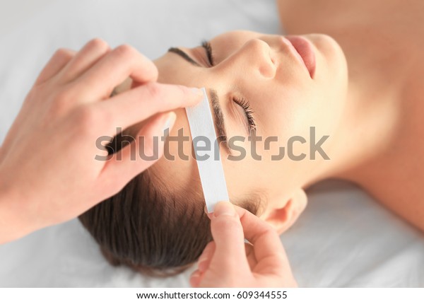 Beautician\
waxing young woman\'s eyebrows in spa\
center